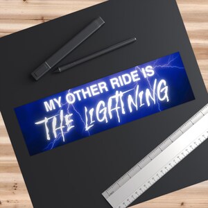 May include: A rectangular bumper sticker with the text "MY OTHER RIDE IS THE LIGHTNING" in white, outlined with a blue glow, set against a blue background with lightning bolts. The sticker is on a black surface.