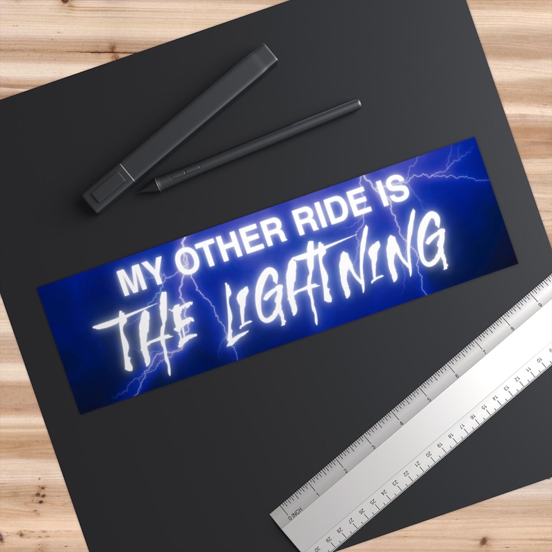 May include: A rectangular bumper sticker with the text "MY OTHER RIDE IS THE LIGHTNING" in white, outlined with a blue glow, set against a blue background with lightning bolts. The sticker is on a black surface.