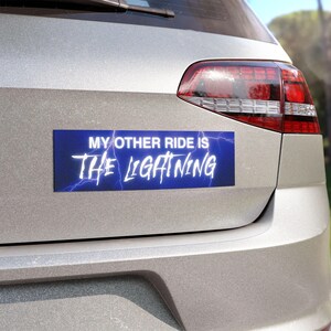 May include: A rectangular blue bumper sticker with the text "MY OTHER RIDE IS THE LIGHTNING" in white, stylized font. Lightning bolt graphics are behind the text. The sticker is affixed to the back of a silver car.