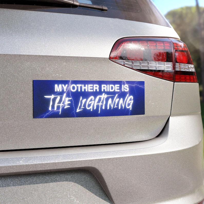 May include: A rectangular blue bumper sticker with the text "MY OTHER RIDE IS THE LIGHTNING" in white, stylized font. Lightning bolt graphics are behind the text. The sticker is affixed to the back of a silver car.