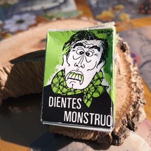 Puede incluir: Una caja rectangular con una ilustración gráfica en verde y negro de una persona con expresión de sorpresa que lleva una bufanda de cuadros verdes. El texto de la caja dice Broma y Dientes Monstruo en letras blancas y negras.