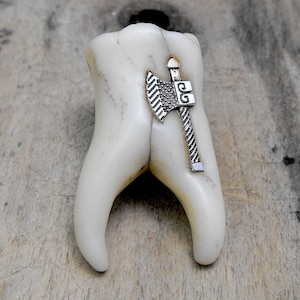 May include: A white, tooth-shaped object featuring a silver-toned metal axe charm attached to its surface. The axe charm has a textured blade and a detailed handle, positioned vertically against the smooth, off-white surface of the tooth.