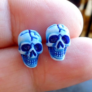 May include: Two small, three-dimensional skull beads featuring a white base color with deep blue detailing in the eye sockets, nasal cavity, and teeth. The surface of each skull bead has a smooth, polished finish with visible carved lines.