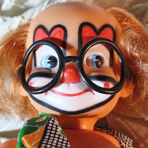 Puede incluir: Una muñeca payaso con piel color melocotón, cabello naranja y ojos azules. El rostro presenta cejas pintadas en forma de corazón rojo, una nariz roja y una amplia sonrisa roja. La muñeca lleva grandes gafas circulares de plástico negro y un cuello de tela con patrones coloridos.