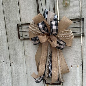 May include: A rustic farmhouse-style bow made of burlap and black and white checkered ribbon. The bow is attached to a metal wall basket. The bow has multiple layers and textures, with a mix of solid and patterned ribbons. The background is a weathered wooden wall.