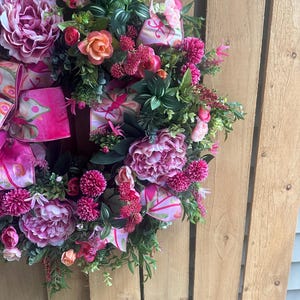 May include: A vibrant floral wreath featuring large pink peonies, smaller pink and orange roses, and various greenery. The wreath is adorned with pink patterned ribbons and is set against a wooden background. A decorative piece for doors or walls.