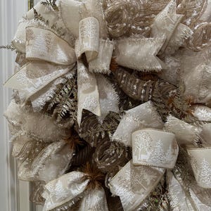 May include: A decorative wreath featuring cream-colored ribbons and accents. The ribbons are adorned with a winter forest pattern. The wreath includes gold and brown mesh and decorative sprigs, creating a festive holiday theme.