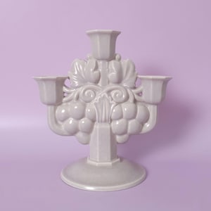 Può includere: Un candelabro in ceramica color lavanda chiaro con tre portacandele. La struttura centrale è decorata con grappoli d'uva in rilievo, motivi a vite vorticosi e disegni di foglie. Poggia su una base circolare con una finitura liscia e lucida.
