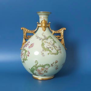 Puede incluir: Un jarrón de cerámica verde pálido con asas de metal dorado ornamentadas y un borde dorado. El cuerpo del jarrón está decorado con delicados patrones florales en tonos rosa, morado y verde, acentuados por intrincados diseños de volutas.