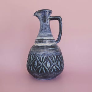 Può includere: Brocca in ceramica blu e nera con manico. La brocca presenta una superficie strutturata, strisce orizzontali bianche e un motivo geometrico. Lo sfondo è rosa.