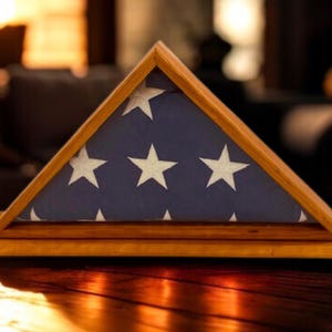 May include: A wooden triangular display case holding a folded American flag. The flag has a blue field with white stars and red and white stripes. The case is made of light-colored wood and sits on a dark surface.