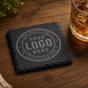 May include: A square black slate coaster with rough, natural edges. The centre features a circular engraved design with the text "YOUR LOGO HERE" surrounded by the repeated phrase "YOUR DESIGN HERE" and small star icons.
