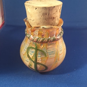 Pode incluir: Uma bolsa de dinheiro de vidro com uma rolha de cortiça. A bolsa é decorada com um sinal de dólar e tem um padrão de espiral multicolorido em tons de laranja, amarelo, verde e azul.