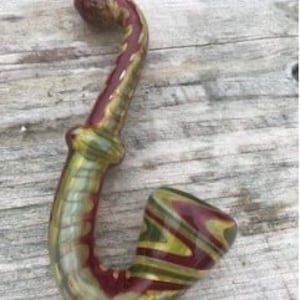 Peut inclure: Une pipe en verre avec un motif en spirale rouge, vert et or. La pipe a un bol rond et une tige incurvée.
