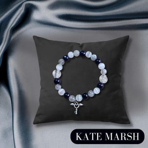 May include: A bracelet with white, navy, and clear beads, and a silver key charm with wings. The bracelet is displayed on a black cushion. The text "KATE MARSH" is at the bottom of the image.