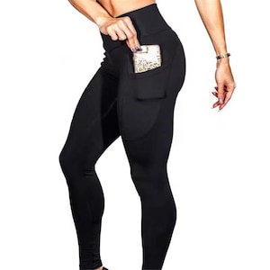 May include: Black leggings with a high waistband and a side pocket. The pocket holds a smartphone with a gold and white patterned case. The leggings are form-fitting and made of a smooth, opaque fabric.