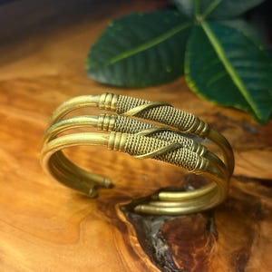 May include: A gold-colored cuff bracelet with a woven design. The bracelet has three bands and a textured, wrapped pattern in the center. The metal has a slightly aged appearance, and the bracelet is set against a wooden surface.
