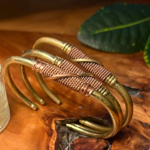 May include: Three brass bracelets with a woven copper and brass design. The bracelets are displayed on a wooden surface. The bracelets have a curved, open design. The woven section is in the center of the bracelet.