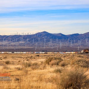 May include: A long freight train travels through a desert landscape with a large wind farm in the background. The train is made up of many boxcars and a locomotive. The wind farm is made up of many wind turbines.