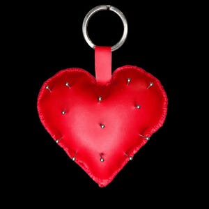 May include: A red heart-shaped keychain with silver pins and a silver key ring. The heart is made of leather and has a small red strap. The background is black.