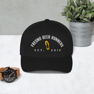Può includere: Cappellino da camionista nero con la scritta "FRESNO BEER RUNNERS" in bianco, arcuata sopra un'impronta di scarpa gialla e oro. La scritta "EST. 2019" è sotto. Un orologio e una piccola pianta sono sullo sfondo.