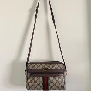 May include: A rectangular crossbody bag featuring the iconic beige and brown GG monogram canvas pattern. The bag includes a brown leather trim, a front zippered pocket with a gold-tone logo emblem, and a central vertical red and green stripe.