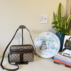 Puede incluir: Bolso bandolera Gucci vintage con el icónico estampado GG, ribetes de cuero marrón y una franja roja y verde. El bolso se exhibe con un letrero que dice "Vintage Re-Vibes by Jelly" y una pila de libros de moda.