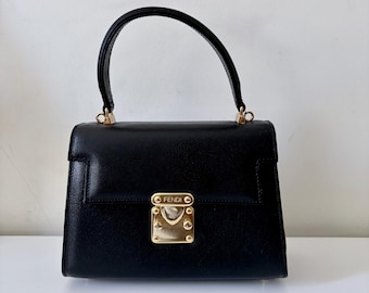 May include: A black leather handbag featuring a single top handle and a gold-tone metal clasp closure on the front flap. The clasp is engraved with the word FENDI and is secured with four small gold-tone rivets. The bag has a structured rectangular shape.