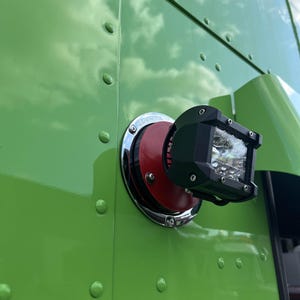 May include: A black and silver LED light mounted on a green surface with a red cap. The light has a clear lens and is surrounded by a black housing. The light is mounted on a green surface with a red cap.