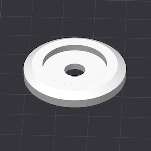 May include: A white, circular object with a raised outer rim and a central hole. The object appears to be made of a solid material and is set against a dark, grid-patterned background. The design is simple and geometric.