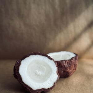 Może przedstawiać: Dwie świece w kształcie kokosa, każda przecięta na pół, leżą na brązowej powierzchni. Zewnętrzna skorupa jest ciemnobrązowa, z białym wnętrzem i knotem. Świece mają przypominać kokos.