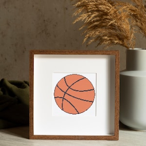 May include: A framed cross-stitch artwork featuring a basketball design. The basketball is orange with black lines, set against a white background. The frame is a warm brown color, and the artwork is displayed near a vase and dried plants.