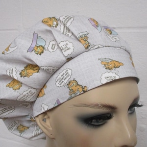 May include: A light gray scrub cap featuring a repeating pattern of the cartoon character Garfield in various poses. Speech bubbles with text are also included in the design. The cap is made of a soft fabric.