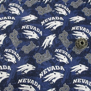 May include: Navy blue fabric with a repeating pattern of the word "NEVADA", the word "Pack", and a white wolf graphic. A metal bobbin sits on the fabric. The fabric has a mottled appearance.
