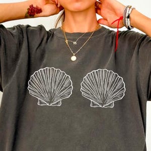 May include: A charcoal gray short-sleeve t-shirt featuring two white line-art seashell graphics centered on the chest. The shirt has a crew neckline and a relaxed, loose-fitting silhouette with a faded, washed-out fabric appearance.