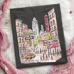 May include: A dark gray t-shirt featuring a whimsical line-art illustration of a New York City street scene. The graphic includes tall buildings, a yellow taxi, pedestrians, and signs reading Broadway, One Way, Hotel, and New York in soft pastel colors.