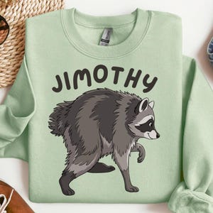 Könnte beinhalten: Ein hellgrünes Sweatshirt mit Rundhalsausschnitt und einer grafischen Illustration eines grauen Waschbären im Profil. Über dem Waschbären ist der Text JIMOTHY in einer dunkelbraunen, abgerundeten serifenlosen Schrift gedruckt. Langarm mit Rippkragen.