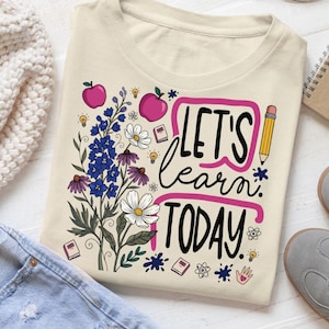May include: A cream-colored t-shirt featuring a graphic print with the text "LET'S learn TODAY." The design includes illustrations of blue and white flowers, two pink apples, a yellow pencil, small books, light bulbs, and atom symbols.