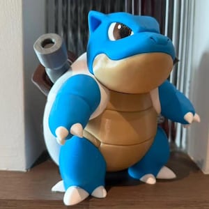 May include: A blue and tan Blastoise figurine featuring a large, rounded head, a tan-colored chest plate, and two prominent gray cannons protruding from the back shell. The character has white claws on its hands and feet and dark eyes.