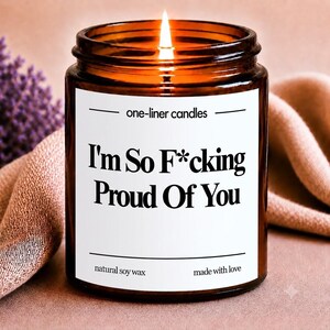 Peut inclure: Une bougie allumée dans un pot en verre ambré avec une étiquette blanche portant le texte one-liner candles, I'm So F*cking Proud Of You, natural soy wax, et made with love. La bougie a une flamme visible et contient de la cire de soja.