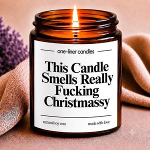 Peut inclure: Une bougie allumée dans un pot en verre brun avec une étiquette blanche portant le texte noir : one-liner candles, This Candle Smells Really Fucking Christmassy, natural soy wax, made with love. La bougie a une mèche allumée.