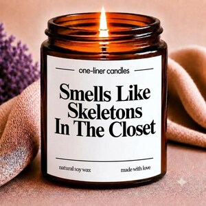 Peut inclure: Une bougie allumée dans un pot en verre ambré avec une étiquette blanche portant le texte one-liner candles, Smells Like Skeletons In The Closet, natural soy wax et made with love. La bougie a une mèche allumée et une flamme visible.