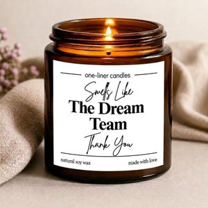 May include: A brown glass jar candle with a white label featuring the text one-liner candles Smells Like The Dream Team Thank You natural soy wax made with love. The candle has a visible lit wick and is filled with wax inside the amber container.
