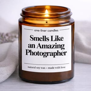 Op de afbeelding: Een brandende kaars in een bruine glazen pot met een wit etiket. Op het etiket staat "Smells Like an Amazing Photographer". De kaars is gemaakt van natuurlijke sojawas en is een one-liner kaars.