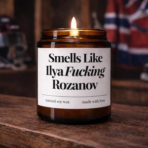May include: A lit brown glass jar candle with a white label that reads "Smells Like Ilya Fucking Rozanov". The candle is made with natural soy wax and made with love.