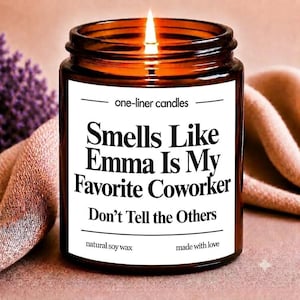 Peut inclure: Une bougie allumée dans un pot en verre ambré avec une étiquette blanche portant le texte noir : one-liner candles, Smells Like Emma Is My Favorite Coworker, Don't Tell the Others, natural soy wax, et made with love.