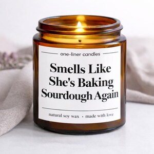 May include: A lit, amber-coloured glass jar candle with a white label. The label reads "Smells Like She's Baking Sourdough Again" in black text. The candle is made with natural soy wax and is made with love.