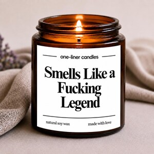 Peut inclure: Une bougie allumée dans un pot en verre brun avec une étiquette blanche. L'étiquette indique "Smells Like a Fucking Legend" en gras noir. La bougie est étiquetée "one-liner candles", "natural soy wax" et "made with love".