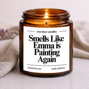 Op de afbeelding: Een brandende sojawas kaars in een bruine glazen pot. Het witte etiket luidt "Smells Like Emma is Painting Again". De kaars is gelabeld met "one-liner candles" en "made with love".