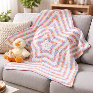 Puede incluir: Una manta de crochet en forma de estrella, con rayas blancas, rosas, azules y amarillas. La manta está sobre un sofá gris, con un juguete de pato de peluche. El diseño de la manta presenta una estrella central.
